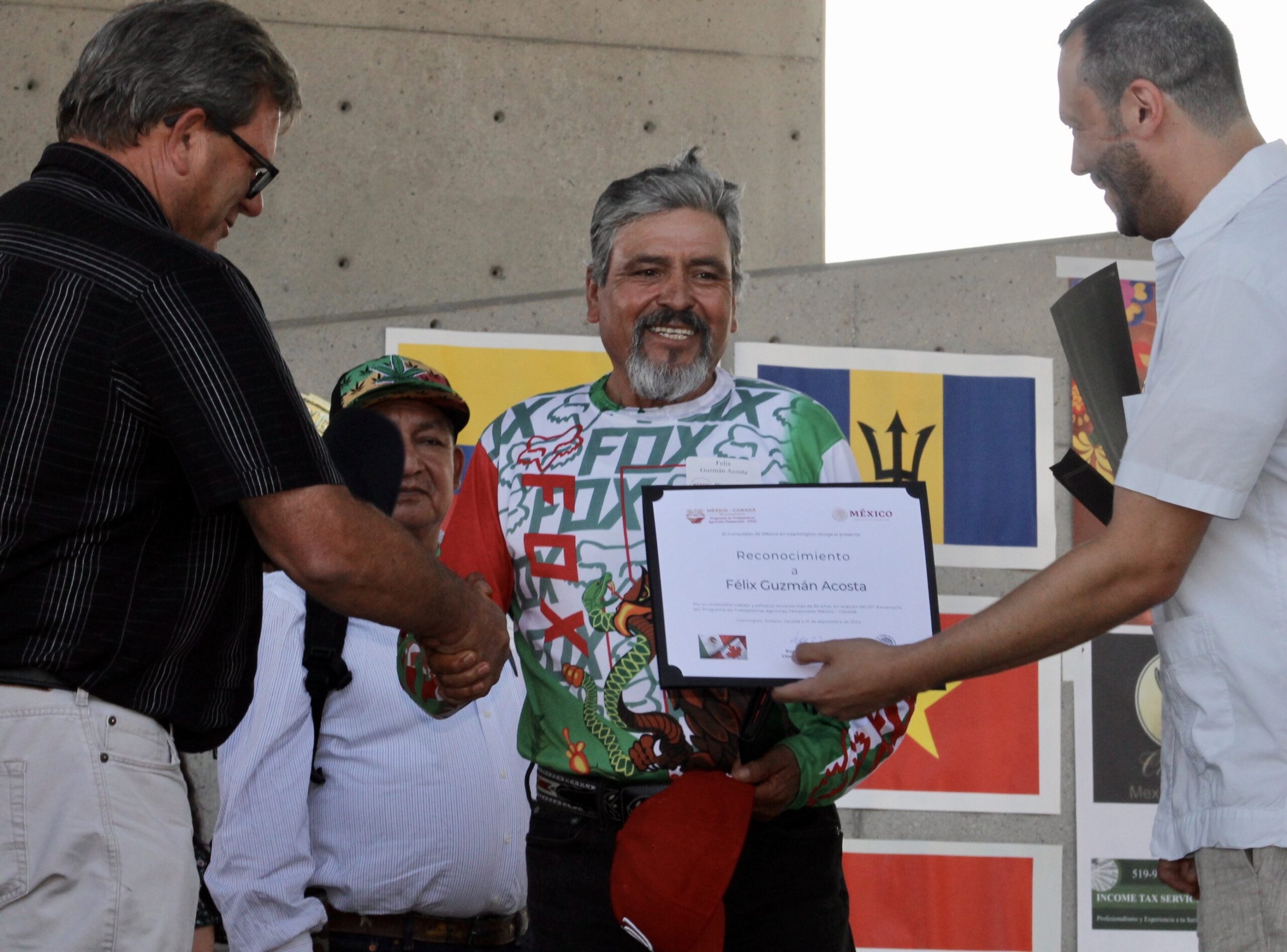 Felix Guzman Acosta, wearing a colorful shirt with a dragon design, smiles as he holds a certificate. On the left, a man in a black shirt shakes his hand, and on the right, another man in a white shirt presents the certificate. A third man in a cap stands in the background. Flags are displayed on the wall behind them.