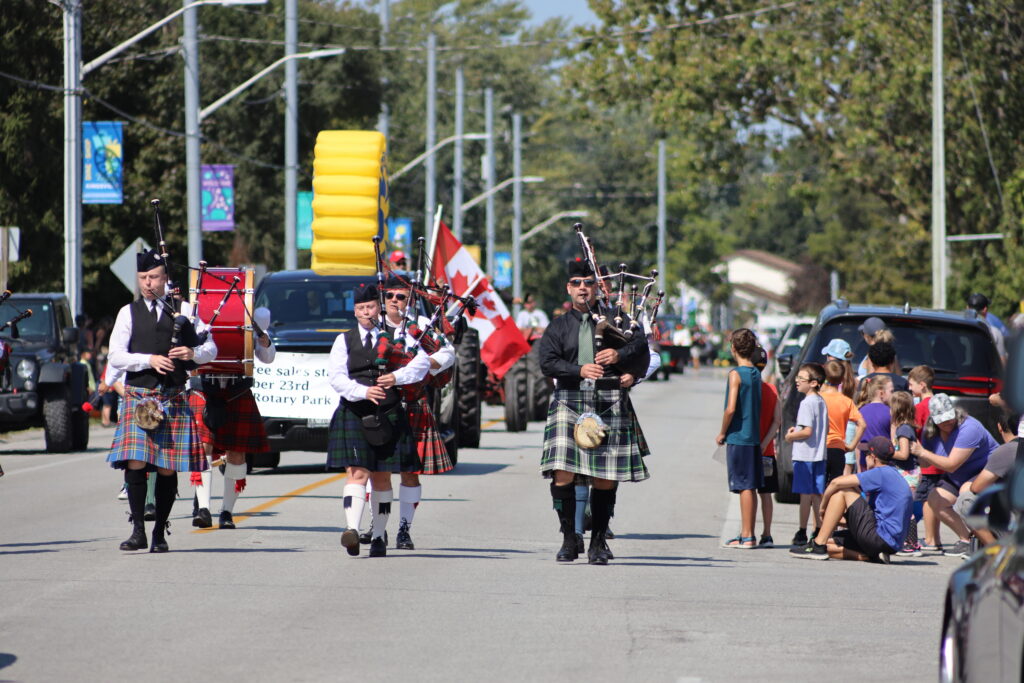 A parade scene on a sunny day features a group of bagpipers in traditional Scottish kilts marching down a street. They are followed by a vehicle with a large inflatable yellow object on top and a Canadian flag. Spectators, including children, line the sides of the street, watching the procession. Trees and parked cars are visible in the background.