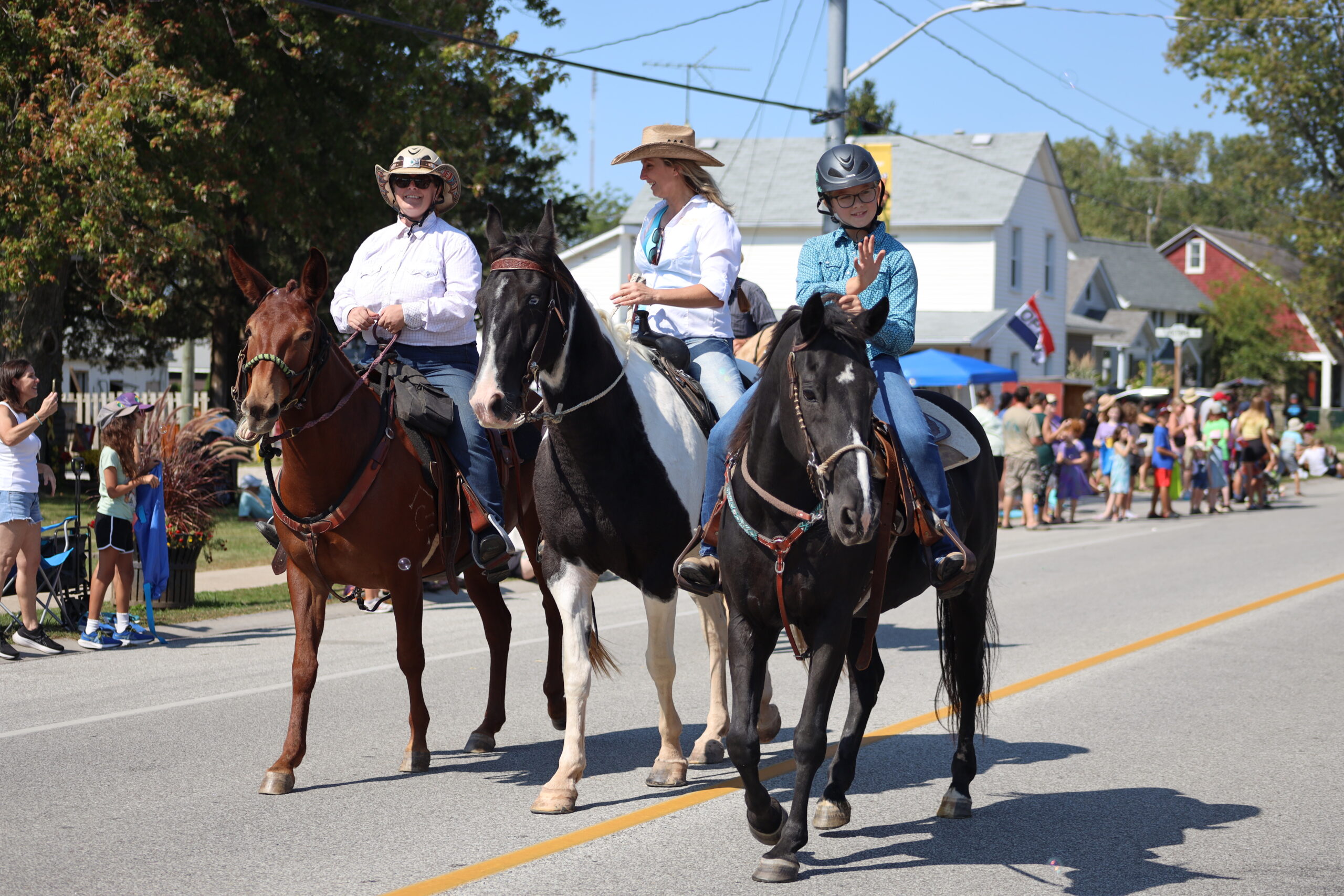 Three people are riding horses down a street during a parade. The person in the middle, wearing a cowboy hat and white shirt, is Sarah Parks. They are flanked by two friends, each on their own horse. The person on the left wears a wide-brimmed hat and sunglasses, while the person on the right wears a helmet and glasses. Spectators line the street, enjoying the sunny day. Trees and houses are visible in the background.