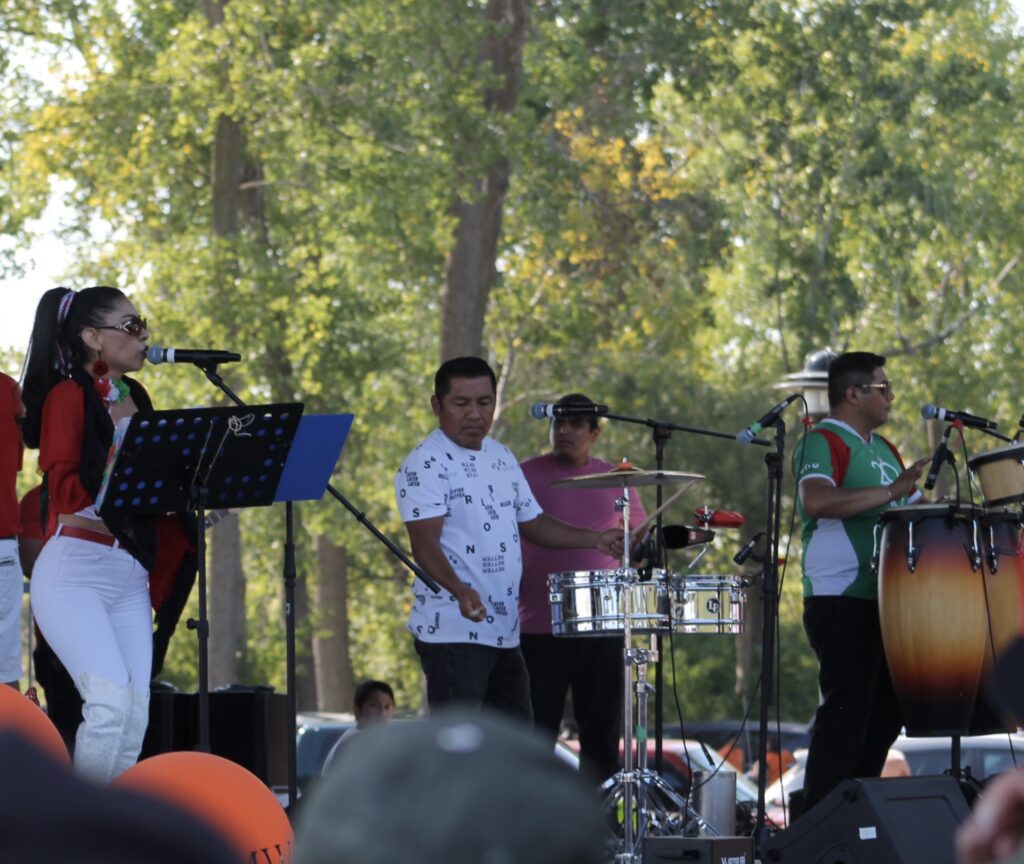 The image captures a lively outdoor musical performance set against a backdrop of lush green trees. A female singer, dressed in vibrant red and white attire, passionately sings into a microphone, while a group of musicians accompanies her. The ensemble includes a percussionist in a white patterned shirt and another musician in a green jersey playing congas. The scene is filled with energy and rhythm, with microphones and instruments set up on stage, suggesting a festive atmosphere. The audience, partially visible, adds to the communal and joyful vibe of the event.