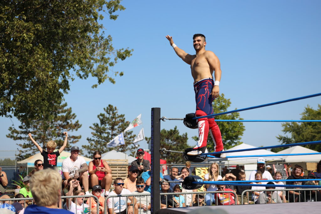 A wrestler stands confidently on the corner ropes of a wrestling ring, wearing red and blue tights with a lightning bolt design. He waves to a cheering crowd seated behind a metal barrier, with trees and a clear blue sky in the background. The audience includes people of various ages, some capturing the moment with their phones.