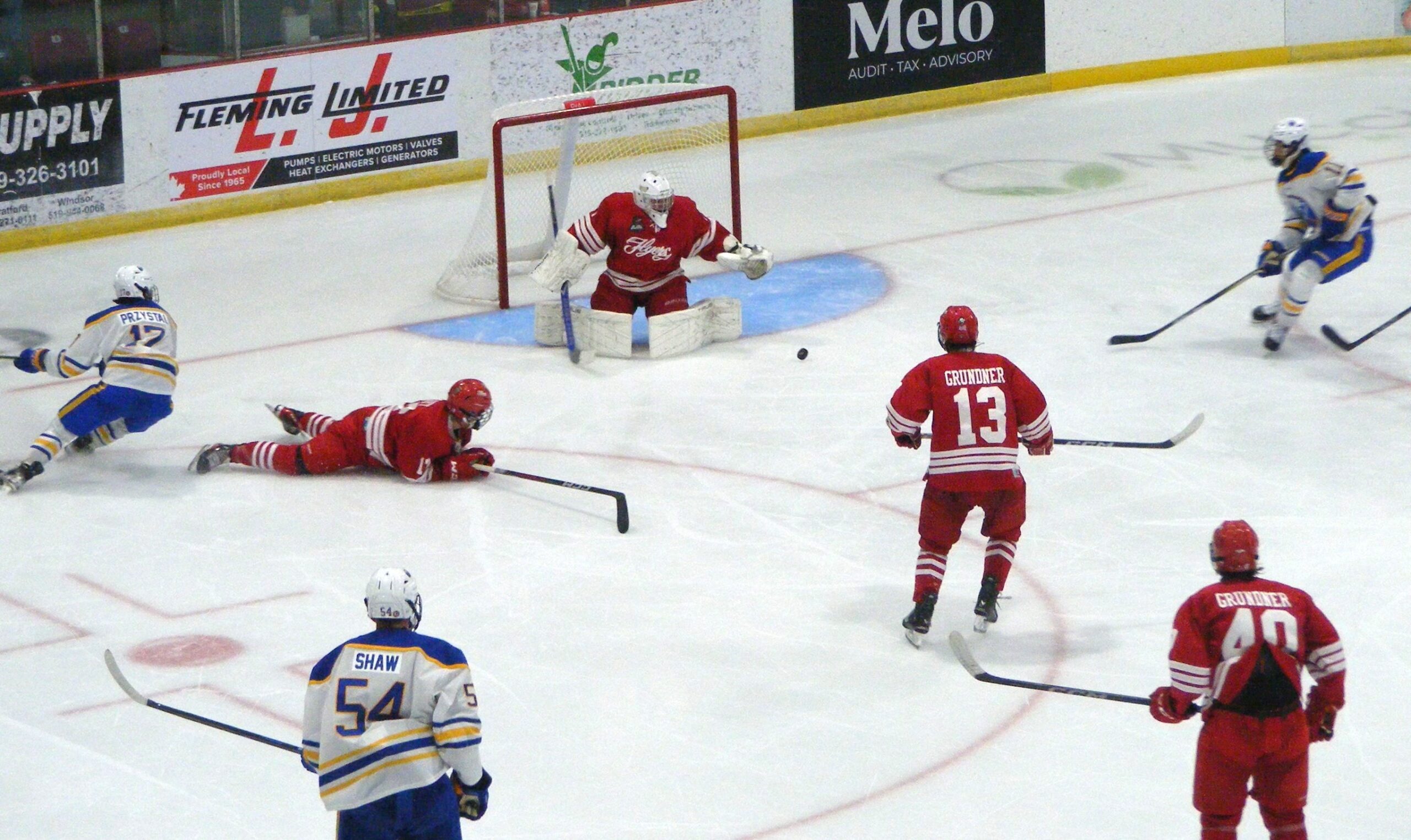 In this dynamic ice hockey scene, players clad in vibrant red and blue uniforms are captured in the midst of an intense game. The red team's goalie, positioned in front of the net, is poised to block an incoming puck, while a player in red lies on the ice, having just attempted a defensive maneuver. The opposing team, dressed in blue and yellow, is actively engaged, with one player sliding across the ice and another preparing to make a play. The rink is surrounded by advertisements, adding to the authentic atmosphere of a competitive sports arena.