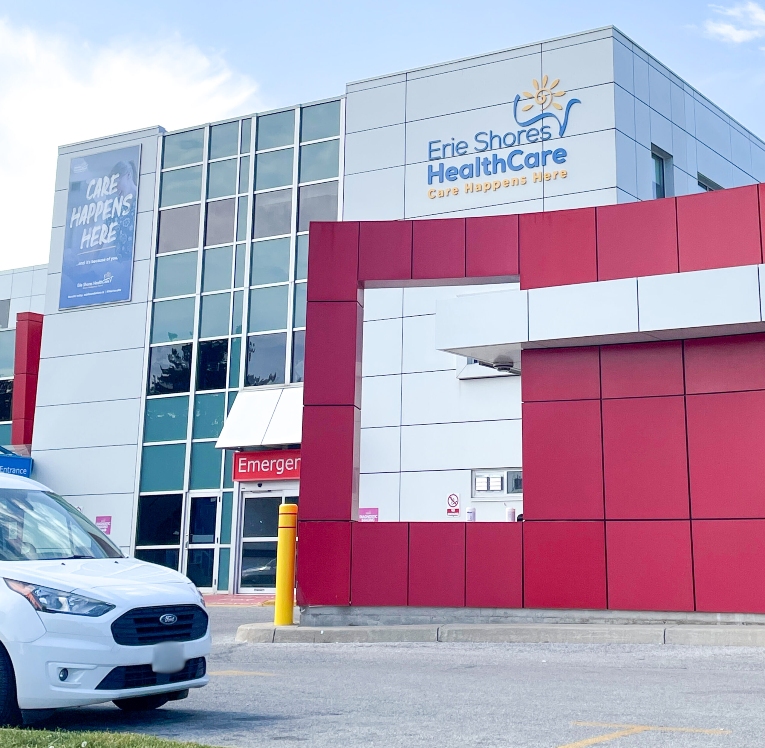 The image depicts a modern healthcare facility with a sleek, contemporary design. The building features a prominent red and white color scheme, with large glass windows reflecting the sky. The signage reads "Erie Shores HealthCare" with the tagline "Care Happens Here," emphasizing its role as a medical center. A white vehicle is parked in front, and the entrance to the emergency department is visible, marked by a red sign. The overall atmosphere is clean and professional, conveying a sense of accessibility and readiness.