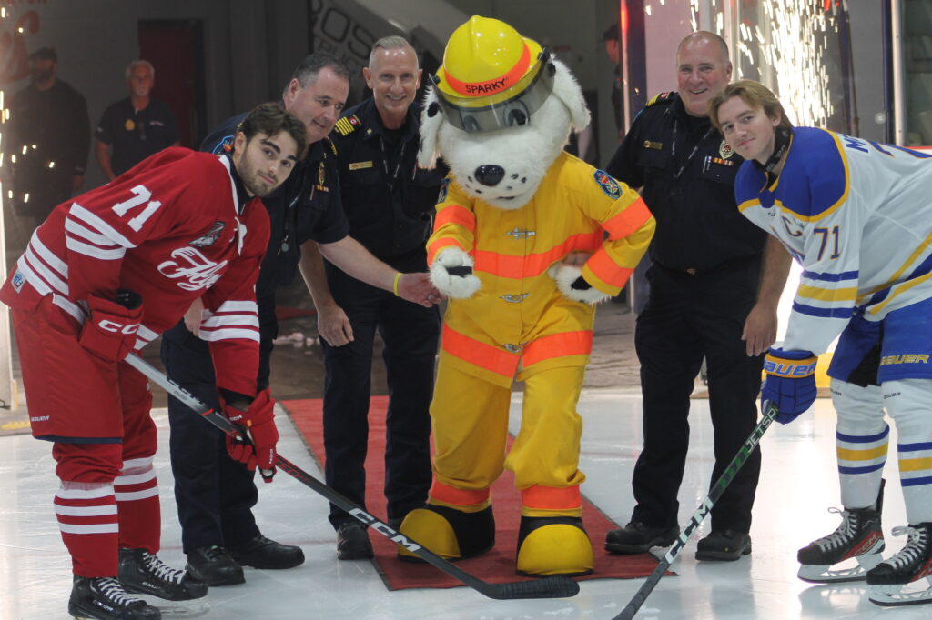 The image captures a lively moment on an ice rink, featuring two hockey players in vibrant uniforms—one in red and the other in blue and white—poised for a face-off. They are flanked by a group of smiling firefighters in uniform, adding a sense of camaraderie and community spirit. At the center, a playful mascot dressed as a firefighter, complete with a yellow suit and helmet, adds a whimsical touch. The scene is illuminated by sparkling lights in the background, enhancing the festive and celebratory atmosphere.