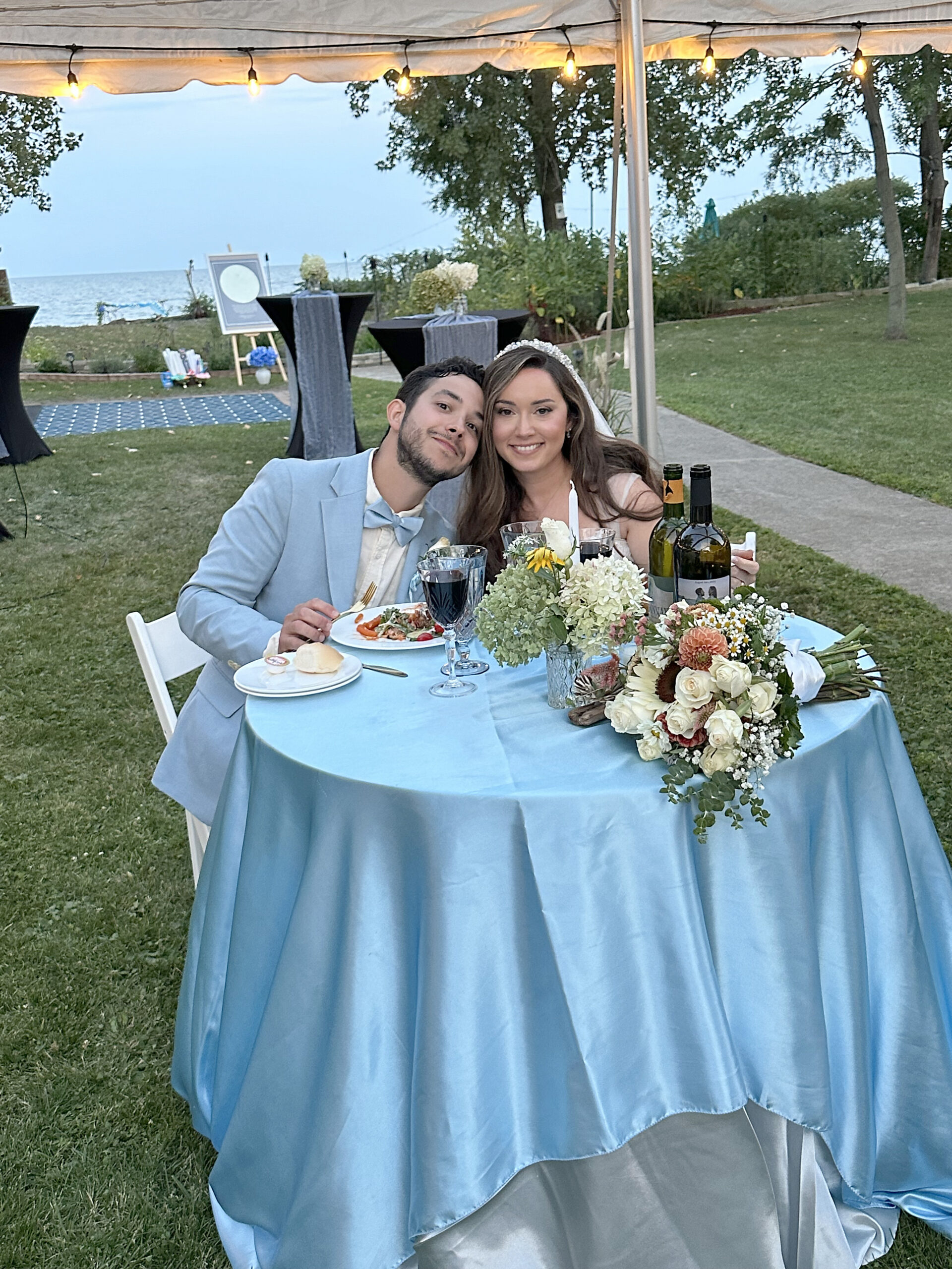 A joyful couple sits at a round table draped in a soft blue tablecloth, set outdoors under a canopy adorned with string lights. The scene is serene, with a backdrop of lush greenery and a glimpse of water in the distance. The table is elegantly arranged with a bouquet of white and blush flowers, two bottles of wine, and a plate of food. The couple, dressed in formal attire, exudes happiness, with the man in a light blue suit and the woman wearing a tiara.