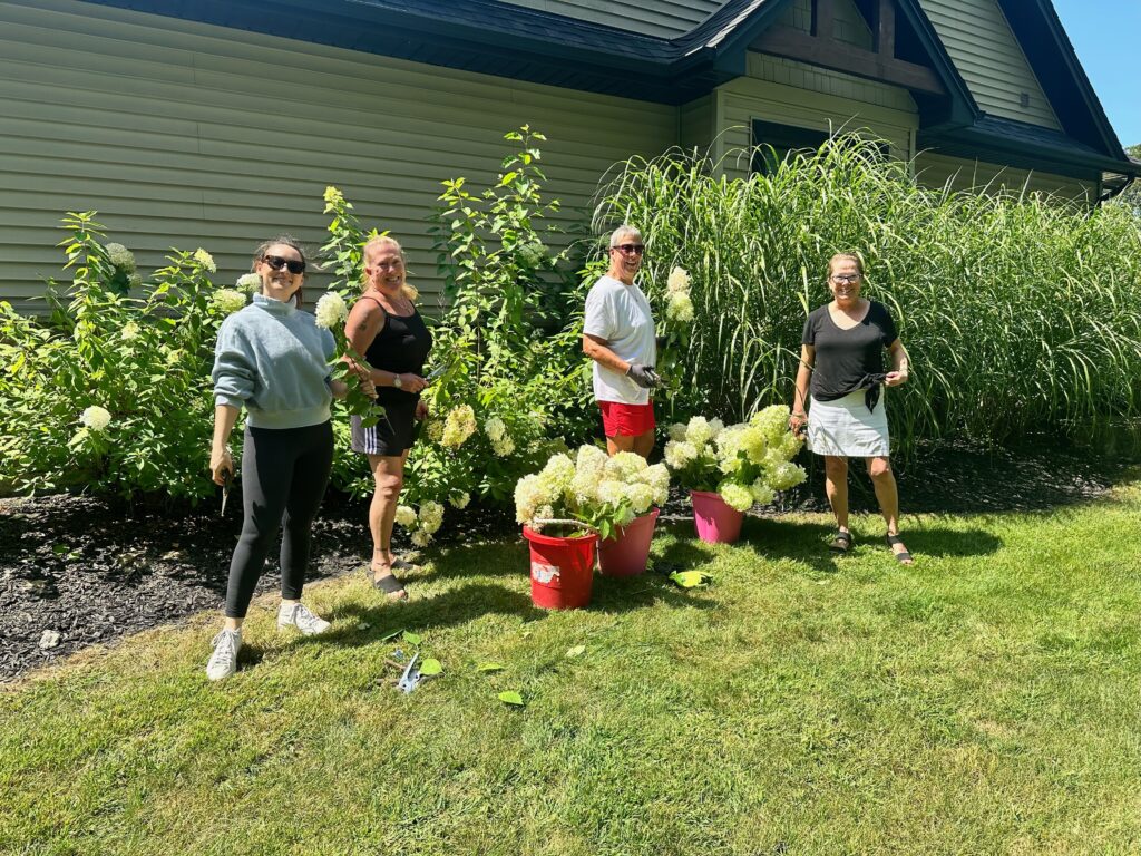 A group of four people stands cheerfully in a lush garden, surrounded by vibrant green foliage and blooming white hydrangeas. They are casually dressed, enjoying a sunny day, with two red buckets filled with freshly cut flowers at their feet. The backdrop features a house with beige siding and a roof with dark shingles, adding a cozy, suburban feel to the scene. The sunlight casts soft shadows, highlighting the joy and camaraderie of the moment as they engage in gardening activities.