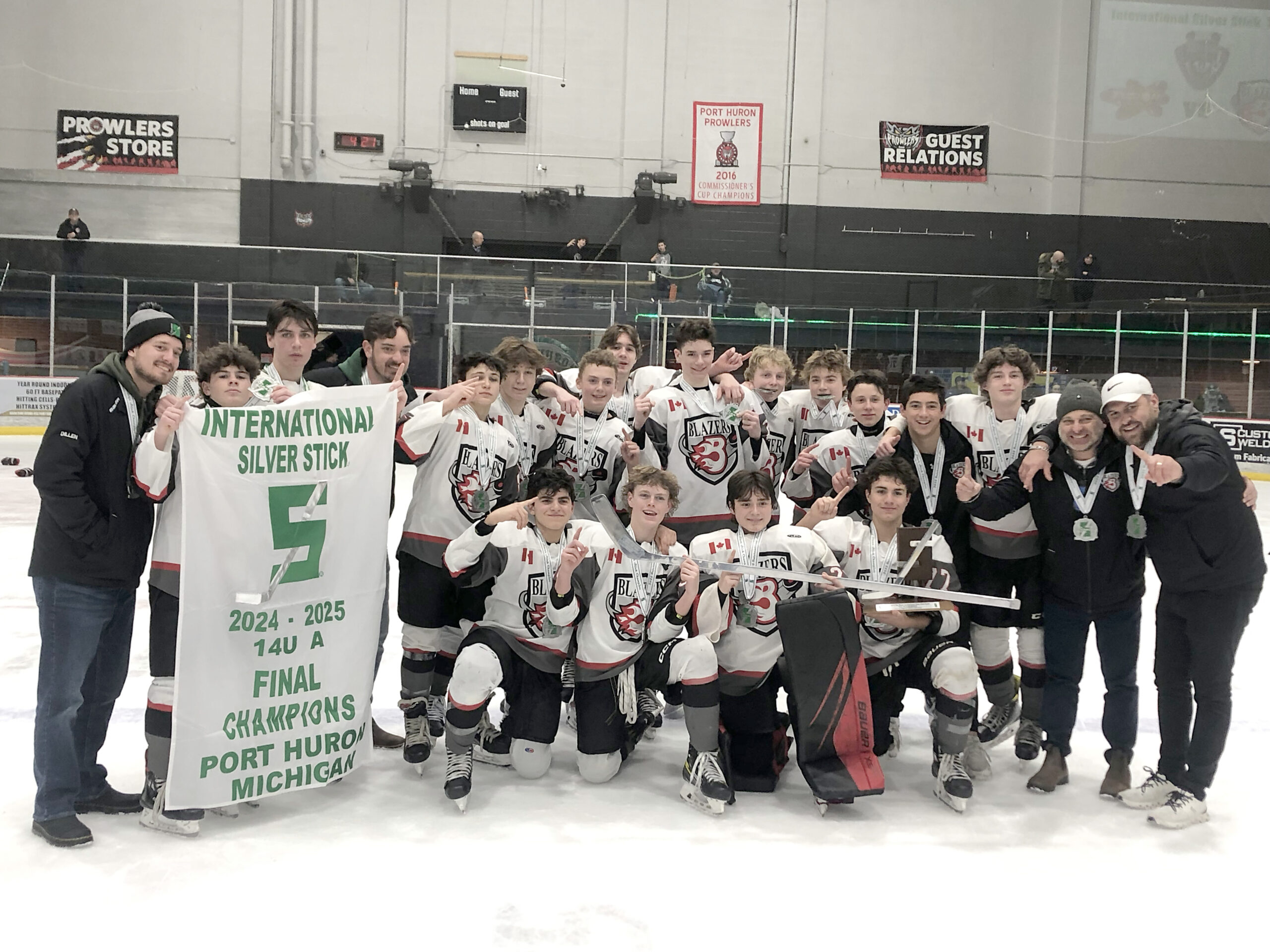 A group of young hockey players in white, red, black and gray uniforms celebrating their victory on the ice. Some players hold a banner that reads "International Silver Stick Final Champions Port Huron Michigan" with additional banners in the background. The players pose for a team photo with a visible trophy in the center.